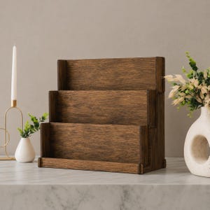 May include: A three-tiered wooden display stand with a dark brown finish and visible wood grain. The structure features three horizontal shelves arranged in a stepped design, providing open storage space for items to be placed upright on each level.