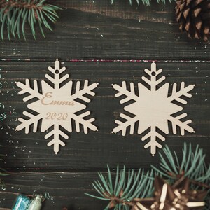 May include: Two wooden snowflake ornaments on a dark wood background. One snowflake has the name "Emma" and the year "2020" engraved on it.
