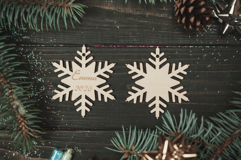May include: Two wooden snowflake ornaments on a dark wood background. One snowflake has the name "Emma" and the year "2020" engraved on it.