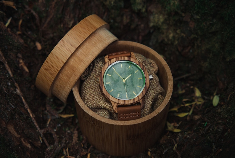 May include: A wooden wristwatch with a green face and brown leather band is displayed in a bamboo box.
