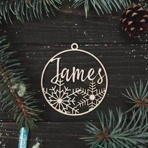 May include: A wooden Christmas ornament with the name "James" cut out in a script font. The ornament is decorated with snowflakes and has a hole at the top for hanging.