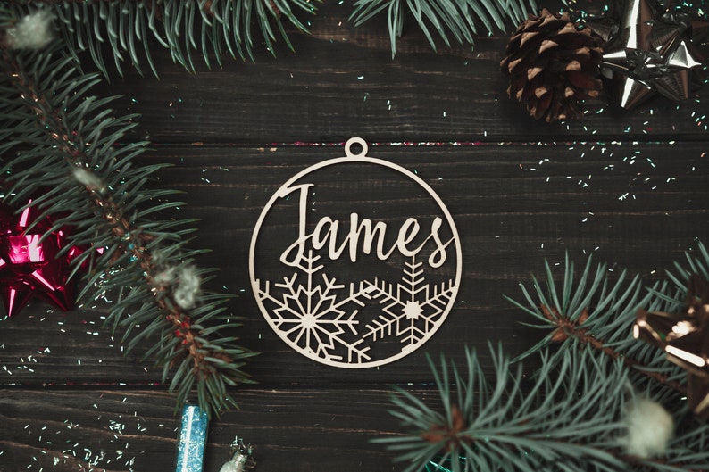 May include: A wooden Christmas ornament with the name "James" cut out in a script font. The ornament is decorated with snowflakes and has a hole at the top for hanging.