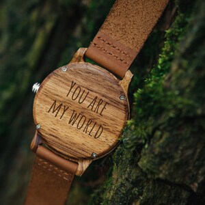May include: A wooden wristwatch with a brown leather band. The watch face is made of wood and has the words "You are my world" engraved on it.