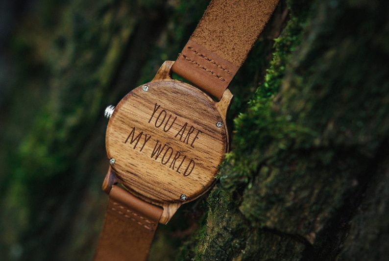 May include: A wooden wristwatch with a brown leather band. The watch face is made of wood and has the words "You are my world" engraved on it.