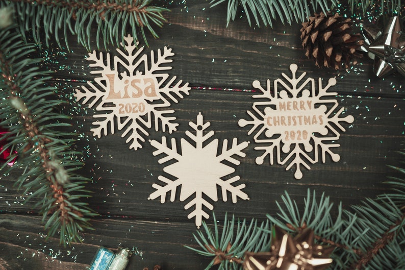 May include: Three wooden snowflake ornaments with a rustic brown finish. One snowflake has the text "Lisa 2020" on it, another has the text "Merry Christmas 2020" on it, and the third is blank.