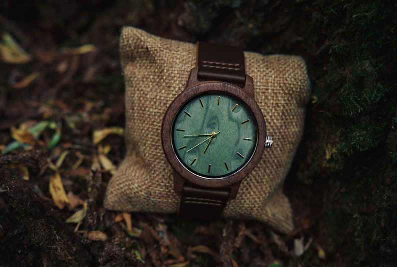 May include: A brown wooden wristwatch with a green wood face and gold hands. The watch is sitting on a brown burlap pouch.