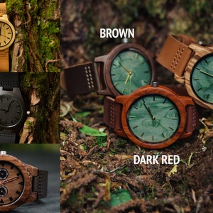 May include: A collection of wooden wristwatches with different colored bands and faces. The watches are arranged on a bed of leaves and twigs. The colors of the watches include light brown, brown, dark red, old black, and new black.