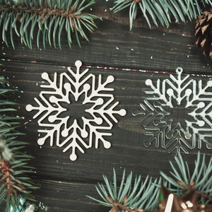 May include: Two white snowflake ornaments on a dark wood background with pine branches and glitter.