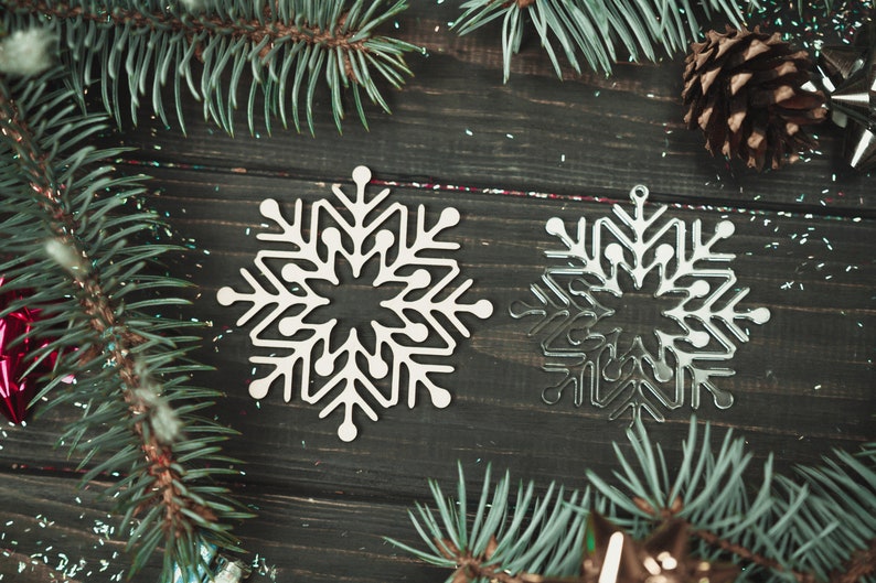 May include: Two white snowflake ornaments on a dark wood background with pine branches and glitter.