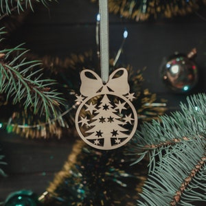 May include: A wooden Christmas tree ornament with a cutout design of a tree and stars. The ornament is hanging from a green ribbon and is surrounded by pine branches.