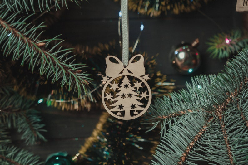 May include: A wooden Christmas tree ornament with a cutout design of a tree and stars. The ornament is hanging from a green ribbon and is surrounded by pine branches.