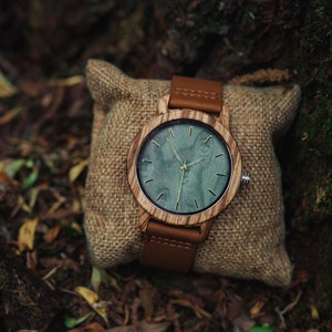 May include: A wooden wristwatch with a brown leather band and a green wood face. The watch is sitting on a brown burlap pouch.