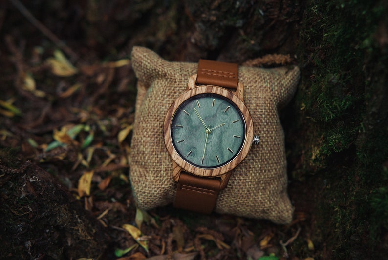 May include: A wooden wristwatch with a brown leather band and a green wood face. The watch is sitting on a brown burlap pouch.