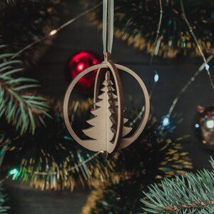 May include: A wooden Christmas tree ornament in the shape of a sphere with a cut-out design of a Christmas tree. The ornament is hanging from a blue ribbon on a Christmas tree.
