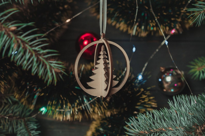 May include: A wooden Christmas tree ornament in the shape of a sphere with a cut-out design of a Christmas tree. The ornament is hanging from a blue ribbon on a Christmas tree.