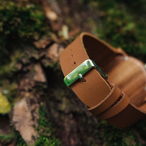 May include: A brown leather watch band with a silver buckle. The band is folded and lying on a mossy surface.