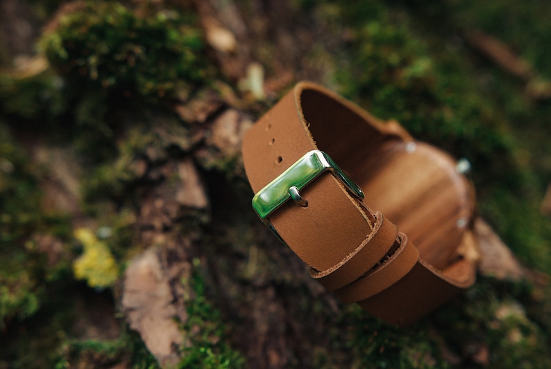 May include: A brown leather watch band with a silver buckle. The band is folded and lying on a mossy surface.
