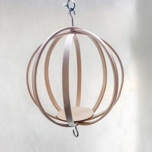 May include: A spherical, wooden hanging planter with a light brown finish. The planter features a series of curved wooden strips forming a cage-like structure, with a circular base inside. A metal hook and chain are attached for hanging.