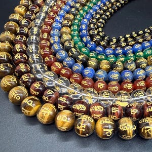 May include: Multiple strands of round beads arranged in a circular pattern. The beads are made of various materials including tiger eye, clear quartz, red, blue, green, and black stones. Each bead features a gold-colored engraved symbol.