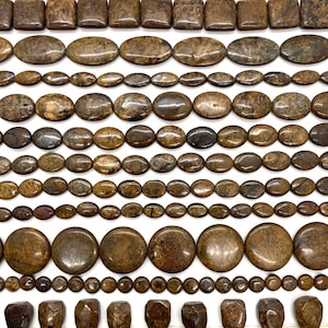 Könnte beinhalten: Eine Sammlung brauner Edelsteinperlen, die in Reihen angeordnet sind und verschiedene Formen wie Quadrate, Ovale, Kreise und facettierte Rechtecke aufweisen. Die Perlen zeigen natürliche, gesprenkelte Muster in dunklen und hellen Brauntönen.