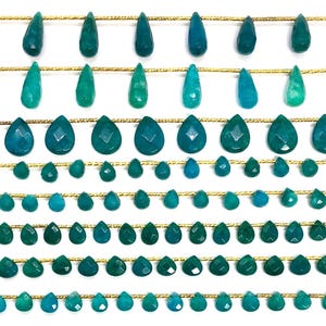 May include: Rows of teal and green gemstone beads in various shapes, including teardrops and faceted drops, strung on gold-colored wire. The beads are arranged in a symmetrical pattern against a white background, showcasing their color variations.