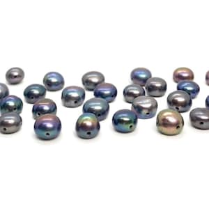 May include: A collection of iridescent, oval-shaped beads. The beads display a range of colors, including blues, purples, and hints of gold. Each bead has a small hole, suggesting they are for stringing. The beads are scattered on a white background.