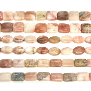 May include: A collection of smooth, polished gemstone beads arranged in six horizontal rows. The beads feature a variety of shapes, including squares, rectangles, and ovals, displaying a natural color palette of soft pink, peach, cream, and grey.