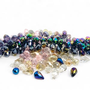 May include: A collection of iridescent and faceted beads in various shapes and sizes. The beads are in shades of purple, blue, green, pink, and clear. Some beads are teardrop-shaped, while others are round or oval. The beads are scattered on a white surface.