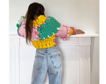May include: A colorful chunky knit sweater with a patchwork design in pink, yellow, green, and white. The sweater has a cropped length and a relaxed fit.