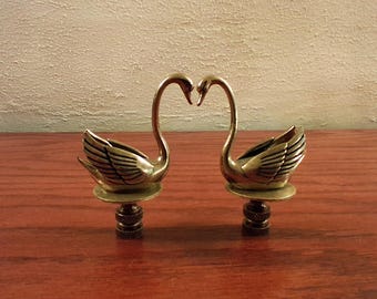 May include: Two brass swan figurines, their necks forming a heart shape. The swans have detailed wings and sit atop a decorative base. The figurines are displayed on a wooden surface.