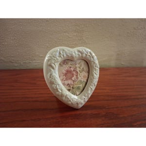 May include: A heart-shaped cream-colored picture frame with a raised floral relief pattern along the border. The center of the frame displays a printed image of a large pink flower surrounded by green leaves and smaller floral accents.