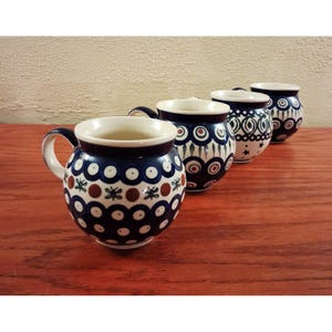 May include: A set of four rounded ceramic mugs featuring a dark blue, white, and brown pattern of circles and small floral motifs. Each mug has a wide, bulbous body and a curved handle, arranged in a diagonal line from the foreground to the back.