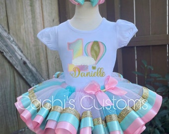 May include: A white t-shirt with a rainbow number one and a hot air balloon design. The shirt says "Danielle" underneath the design. The shirt is paired with a pink and blue tulle tutu with gold glitter ribbon.
