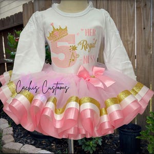 May include: A white long-sleeved shirt and pink tutu dress. The shirt has a gold crown and the number 5 in pink glitter. The tutu has pink, white, and gold ribbons. The text "HER Royal -NESS" is on the shirt.