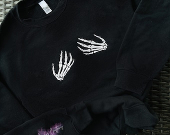 May include: Black sweatshirt with a white skeleton hand design on the front. The sweatshirt has a crew neck and long sleeves.