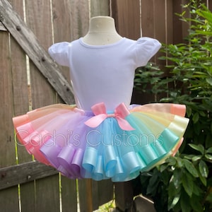 May include: A rainbow tulle tutu skirt with a pink satin bow. The skirt has layers of tulle in shades of pink, yellow, green, blue, and purple. The skirt is worn by a white mannequin.