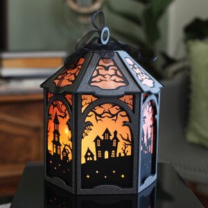 Puede incluir: Una linterna hexagonal hecha de cartulina gris oscuro con purpurina y recortes intrincados de casas encantadas, &aacute;rboles desnudos y murci&eacute;lagos. El interior est&aacute; iluminado con un c&aacute;lido resplandor naranja y un lazo negro en la parte superior.