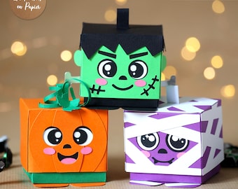 May include: Three paper craft boxes featuring Halloween characters: a green Frankenstein monster, an orange pumpkin, and a purple mummy wrapped in white strips. Each box has large eyes, pink cheeks, and a smiling mouth. Text reads 3D SVG.