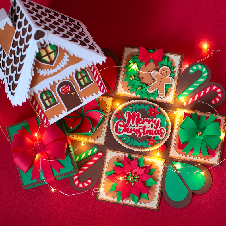 May include: A brown gingerbread house with white icing and red candy hearts sits on top of a multi-layered explosion box. The box is decorated with green, red, and white paper cutouts, including a poinsettia, candy canes, and a Christmas tree. The box also features a red bow, a gingerbread man, and the words "Merry Christmas" in red lettering.