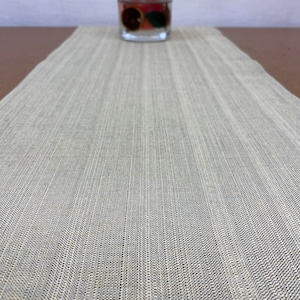 Op de afbeelding: Een beige tafelkleed met een subtiele geweven textuur. De loper is gemaakt van een natuurlijk materiaal en heeft een licht getextureerd oppervlak.