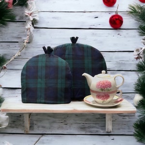 May include: Two green and black plaid tea cozy covers on a white shelf with a white teapot and saucer with pink flowers on it.