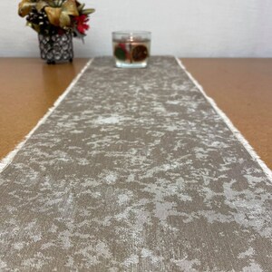 May include: A brown and white patterned table runner with a frayed edge. The runner is made of a linen-like material and has a subtle distressed look.