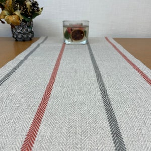 Op de afbeelding: Een grijze en witte gestreepte tafelrunner met rode en grijze strepen. De runner is gemaakt van een geweven stof met een herringbone-patroon.