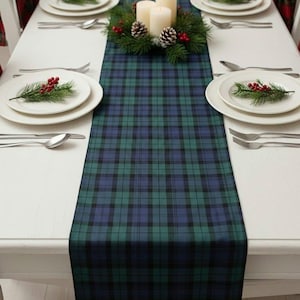 Peut inclure: Un chemin de table rectangulaire avec un motif à carreaux vert foncé et bleu marine. Le chemin est centré sur une table à manger blanche dressée avec des assiettes blanches, des couverts en argent et un centre de table à trois bougies.