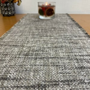 May include: A gray and white woven table runner with a fringed edge. The runner is on a wooden table with a clear glass candle holder in the background.