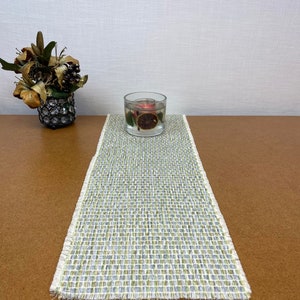 May include: A green and white woven table runner with a frayed edge. A clear glass candle holder with a red candle and dried fruit inside sits on the runner.