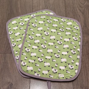 Könnte beinhalten: Ein Paar grüne Ofenhandschuhe mit einem weißen Schaf-Muster. Die Handschuhe haben einen grauen Saum und eine Aufhängeschlaufe.
