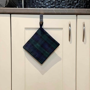 May include: A square, plaid pot holder in navy blue and green hangs from a silver hook on a white cabinet door. The pot holder is diamond-shaped and has a dark blue fabric loop at the top for hanging.