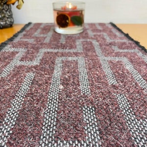 May include: A burgundy and white woven table runner with a geometric pattern. The runner has a fringed edge.
