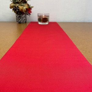 Può includere: Una vivace tovaglia rossa si estende su una superficie di legno. Un piccolo portacandele in vetro con elementi decorativi è posto sulla tovaglia. Un'arrangiamento floreale decorativo in un contenitore di metallo nero è sullo sfondo.